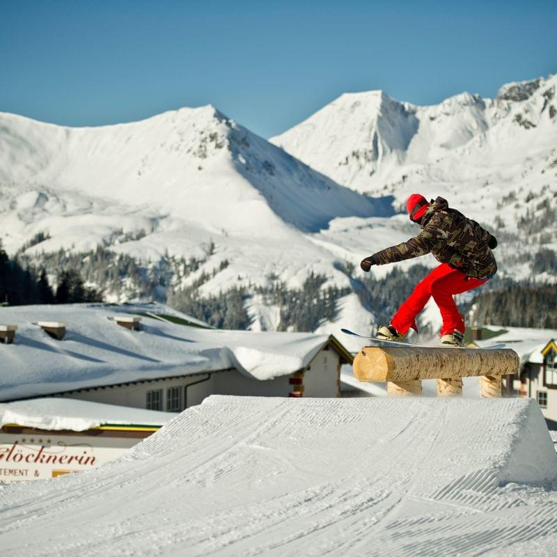 Snowboarder sliding on wooden rail with snowy Alpine mountains in the background